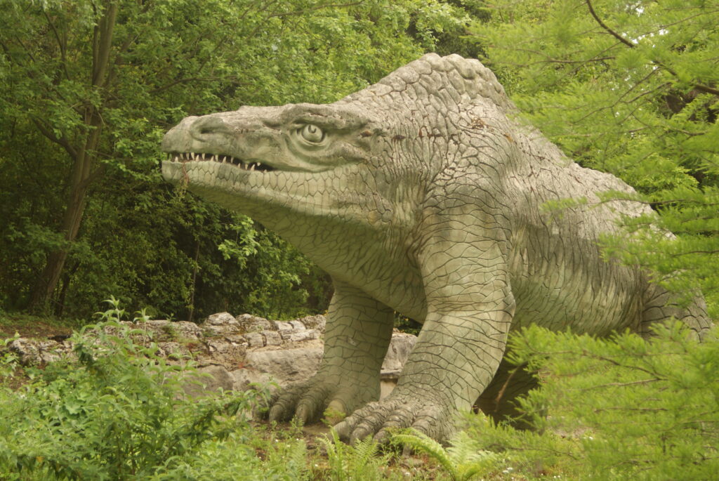 A large, lifelike dinosaur sculpture with textured skin and sharp teeth stands in lush greenery, evoking a prehistoric ambiance in the forest.